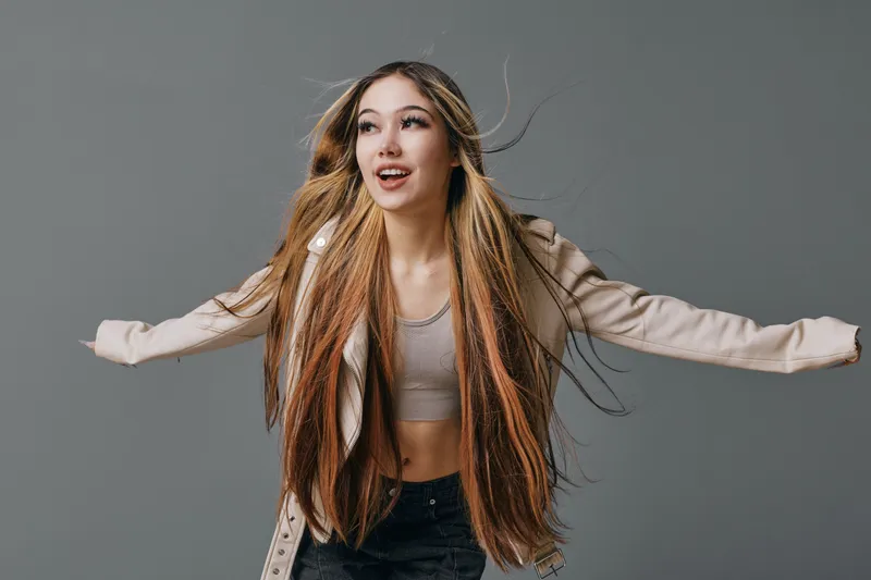 Young woman with long braided hair posing with arms outstretched on gray background wearing casual jacket and crop top for fashion portrait.