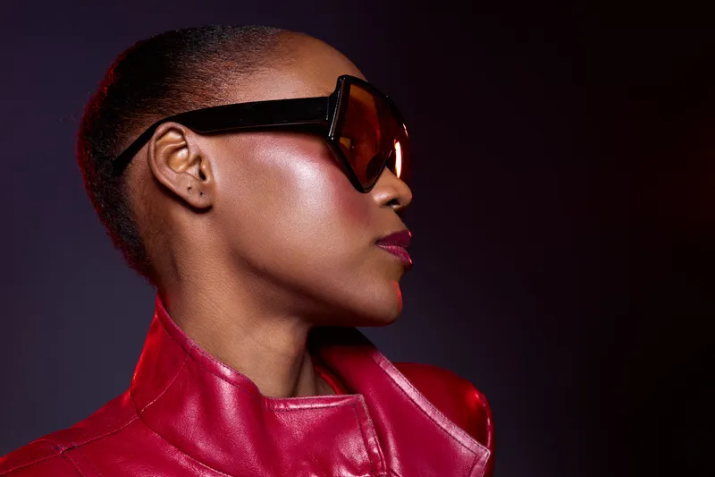 Profile of a confident dark skinned African woman wearing large sunglasses and a red leather jacket against a dark background. Concept of fashion, style, and empowerment.
