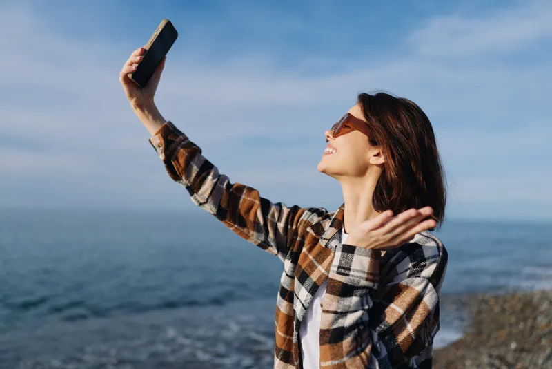 Selfie by the sea with a smiling woman wearing a plaid shirt and sunglasses, enjoying a sunny coastal moment outdoors against a blue horizon in clear weather.