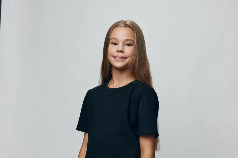 Girl with long hair wearing short sleeve black shirt standing and smiling against light gray background. Portrait of happy child with natural look in studio with calm expression.