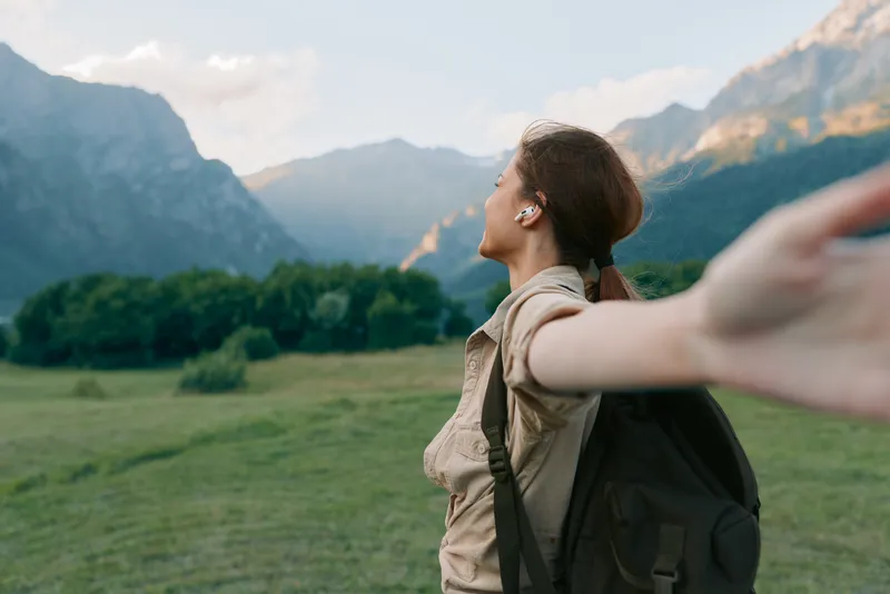 Young woman with a backpack stretches her arms outward in a bright meadow, facing distant mountains to convey freedom, exploration and a deep connection with nature.
