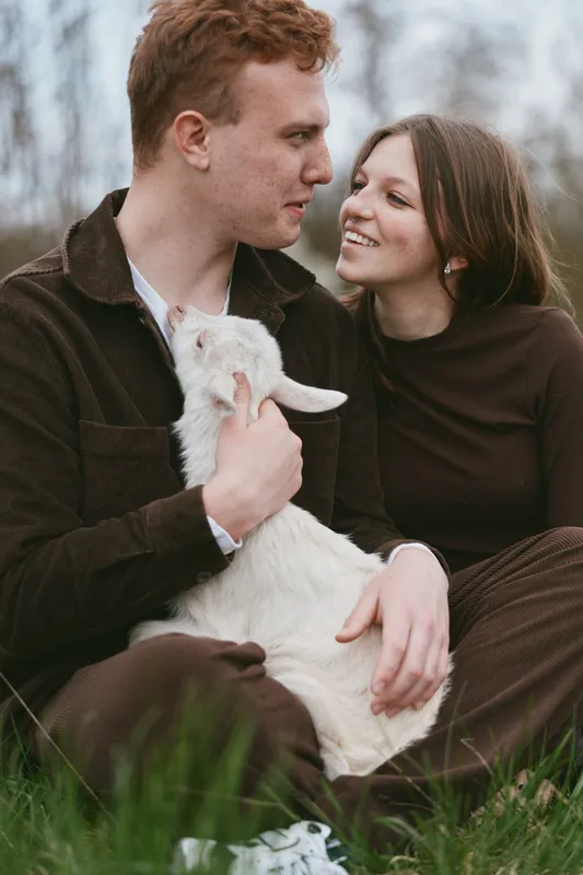 Romantic couple outdoors with a playful goat kid, sharing a tender moment, calm grassy field, warm clothing, soft natural light, candid lifestyle scene, mutual affection
