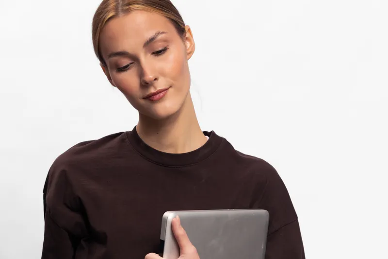 Young woman in dark sweater looks down at tablet with slight smile isolated on white. Concept for technology use, digital lifestyle, and casual modern communication.