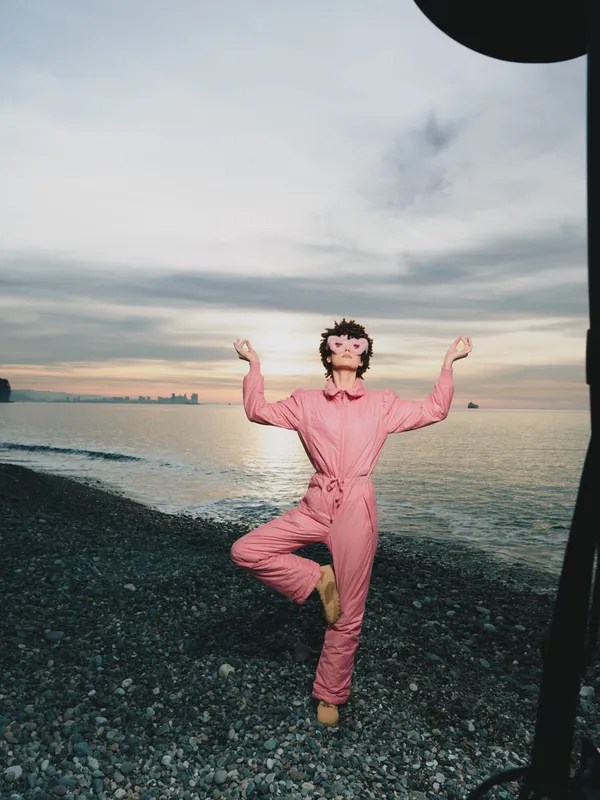 Outdoor fashion on a beach at sunset, a person in a pink jumpsuit performs a balanced pose with arms raised, creating a playful lifestyle moment by the water.