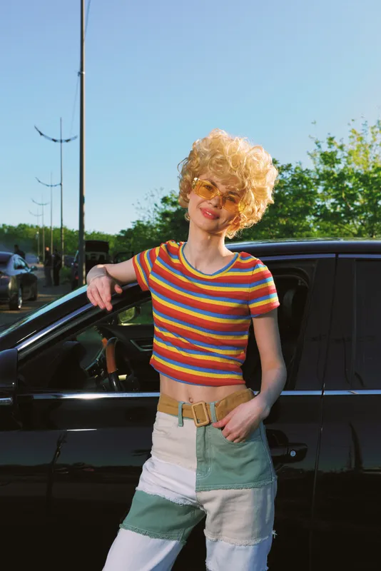 Young woman with curly blonde hair leans on a dark car wearing a bright striped crop top and patchwork jeans, radiating a retro film vibe and carefree confidence.