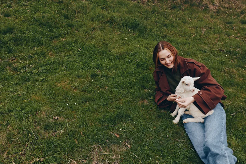 A relaxed woman outdoors sits on the grass, cradling a small white dog and sharing a gentle smile. A lifestyle moment that highlights companionship, outdoor leisure, and calm affection with a beloved
