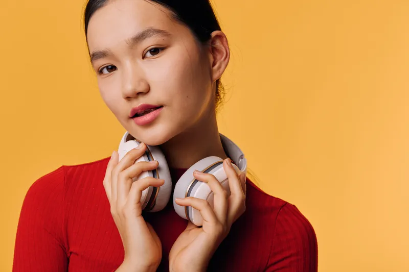 Young woman in red sweater holding headphones close to face with calm expression on orange background, beauty portrait and casual style for music lifestyle or technology concept.