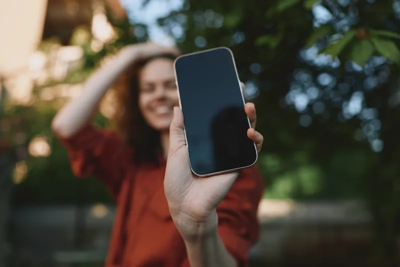 Smiling woman outdoors holding a smartphone toward camera, a clear portrait of modern lifestyle, connectivity, and casual everyday tech. Greenery and daylight create a warm, inviting mood