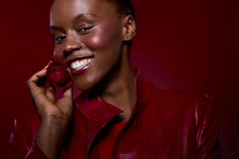 Smiling dark skinned African woman in red leather jacket holds red rose near face on red background. Concept of beauty, fashion, happiness, and natural skin glow in studio portrait.
