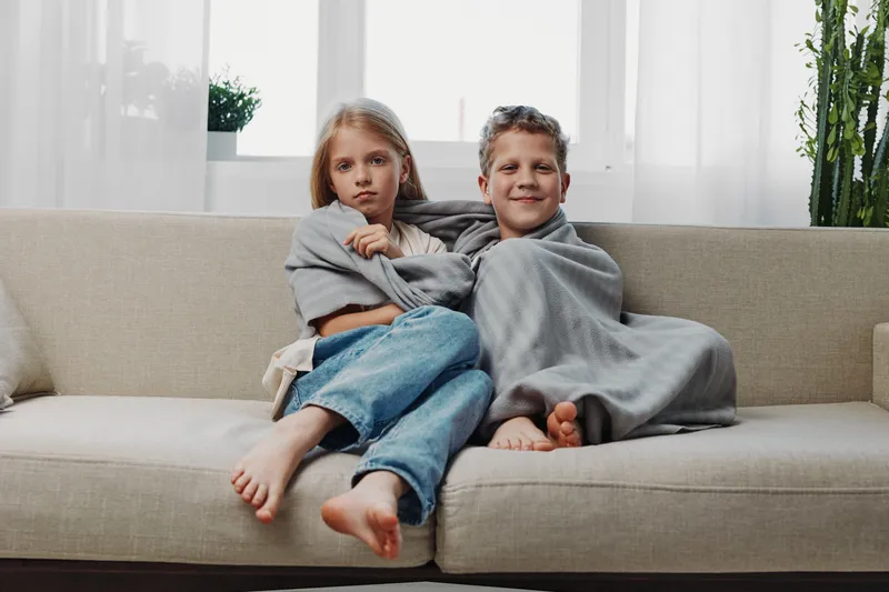 Two children wrapped in grey blanket sit barefoot on sofa in bright living room. The girl looks serious while the boy smiles gently. Concept of sibling bond, comfort, and home warmth.