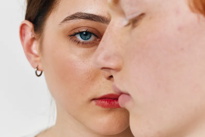 Closeup portrait of two women with clear skin and natural makeup looking calm and serious against a neutral background, focusing on facial features and emotional expression.