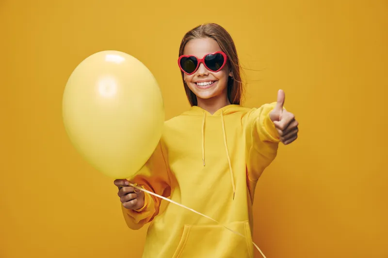 Happy girl in yellow sweatshirt and red heart sunglasses holding balloon with thumb up gesture for positive expression and fun against bright yellow background in studio.