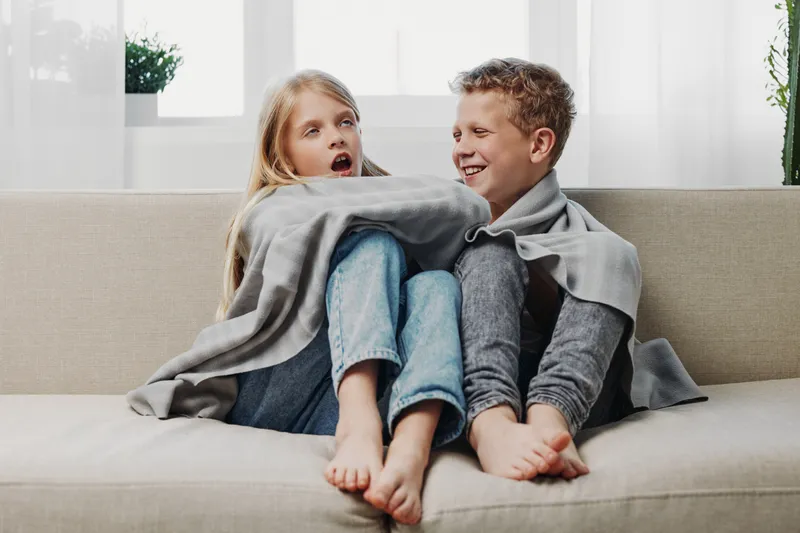 Two children wrapped in a gray blanket sit barefoot on a couch in a bright living room. The girl looks surprised while the boy smiles, creating a cozy and playful home atmosphere.