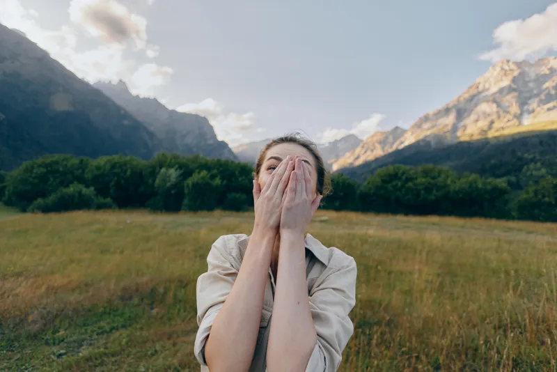 Woman in a light shirt stands in a meadow with distant mountains, hands raised to cover her mouth, expressing playful surprise in a natural outdoor setting.