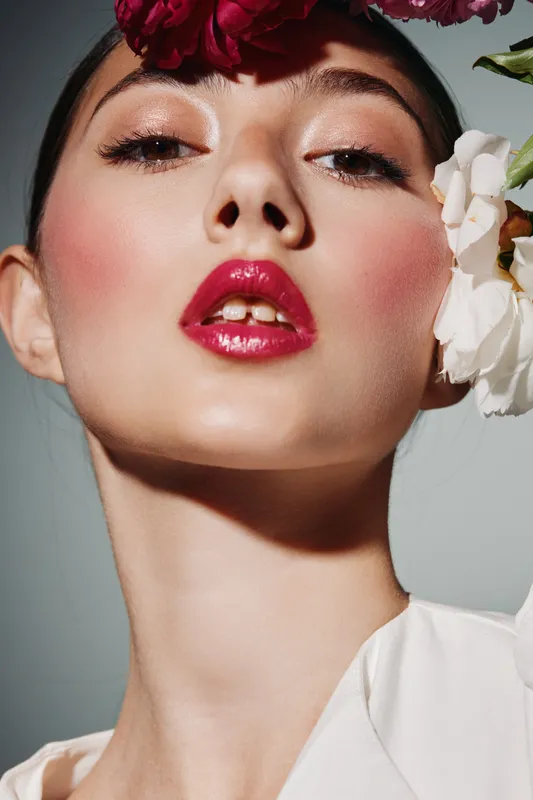 Close-up of a young female model with dramatic makeup and glossy lips posing with flowers on her face and head. Fashion editorial concept with creative beauty styling.