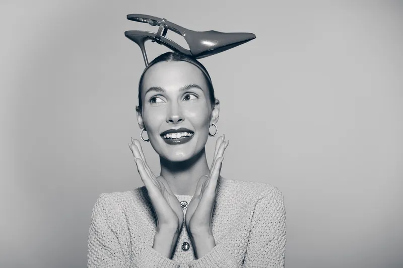 Young white woman with light skin smiling and holding hands near face with shoe on head in studio.