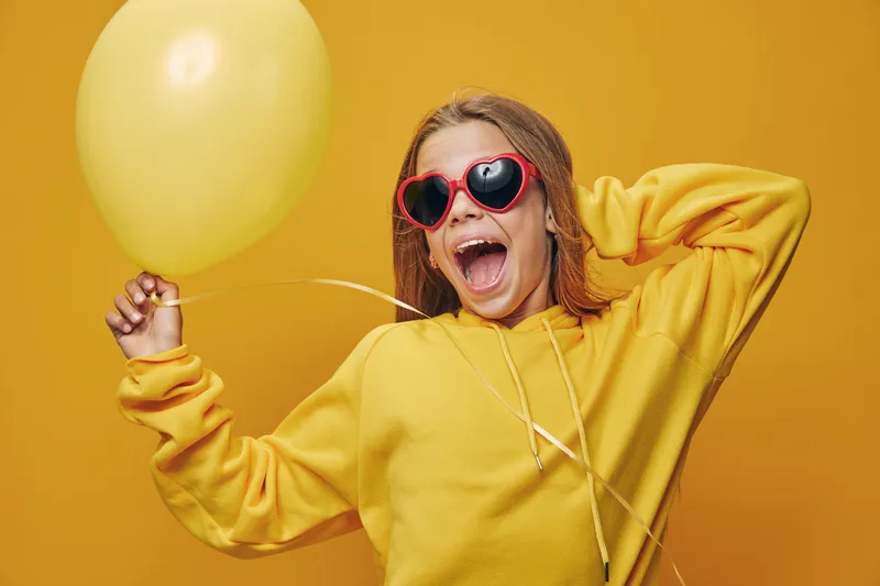Young woman in yellow sweater with red heart-shaped sunglasses holding yellow balloon posing happily on yellow background. Celebration, joy and cheerful mood in portrait.