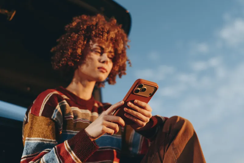 Woman with curly hair holding a smartphone outdoors, casual lifestyle portrait against blue sky. She focuses on the screen, natural light, candid everyday communication moment.