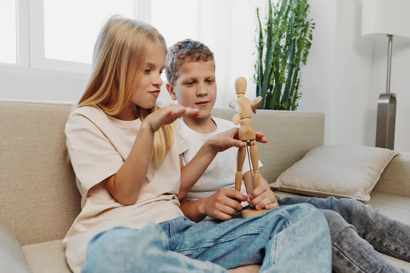 Two children sitting on a sofa playing with an artist mannequin doll, focused and interacting in a bright room with natural light and indoor plants. Concept of creativity and childhood play.