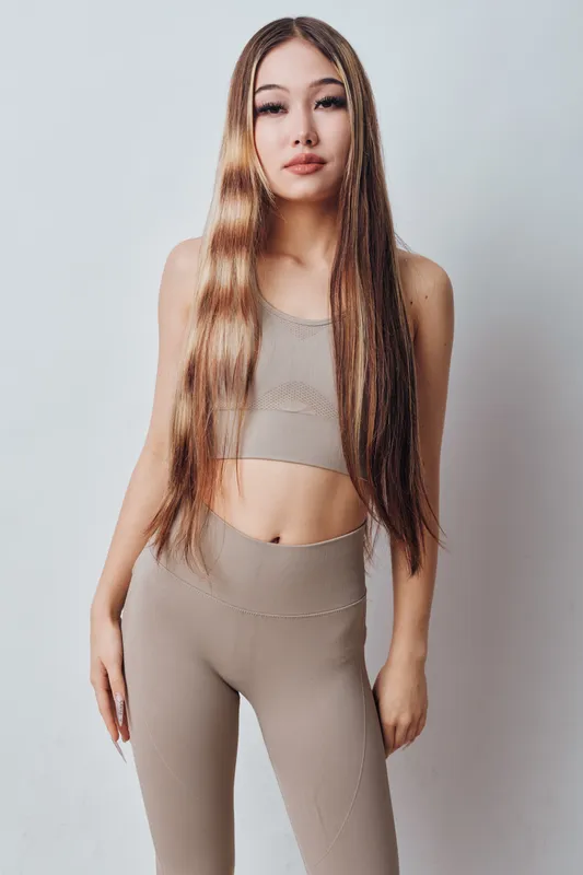 Young woman with long straight hair wearing beige sportswear standing against white background posing confidently for fitness or lifestyle portrait. Slim figure and casual look.