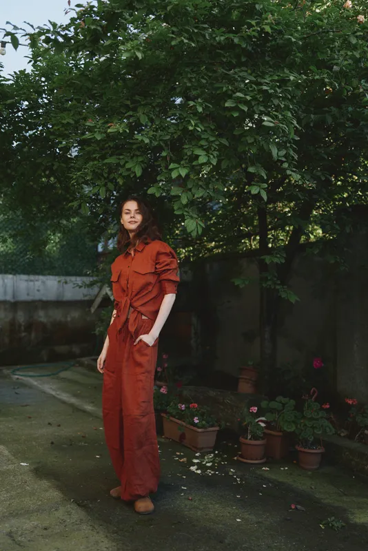 Young woman wearing an orange jumpsuit stands in a garden with potted plants, relaxed pose, natural light, cheerful mood and casual style outdoors