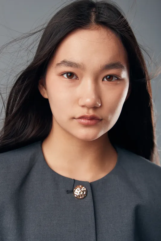 Portrait of young woman with long dark hair and natural makeup wearing gray clothing standing against neutral background with serious expression and direct eye contact.