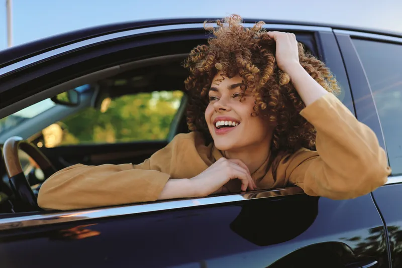 Cheerful woman leaning out of a car window, smiling with sunlit hair, outdoor vibe, casual lifestyle moment, retro film color tones, bright natural hues, candid travel mood