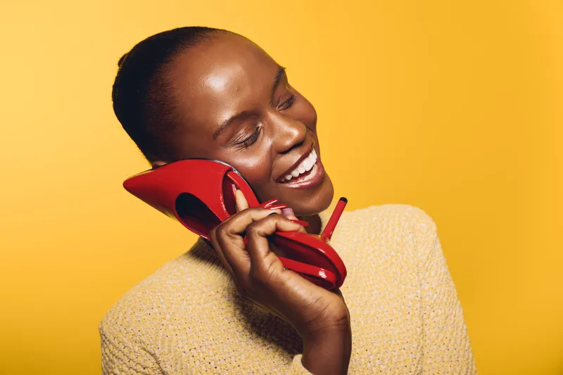Smiling black woman with dark skin holds red high heel shoe to ear like phone, wearing beige sweater against yellow background. Concept of creativity, fun, and fashion expression.
