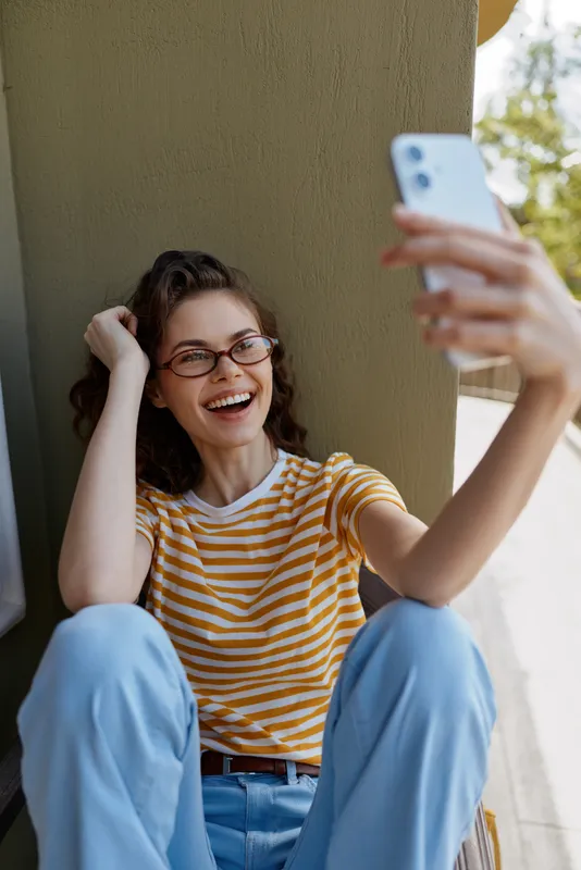 Young woman with brown hair sits against a wall outdoors, taking a selfie with a phone and laughing warmly in a casual striped t shirt and blue jeans.