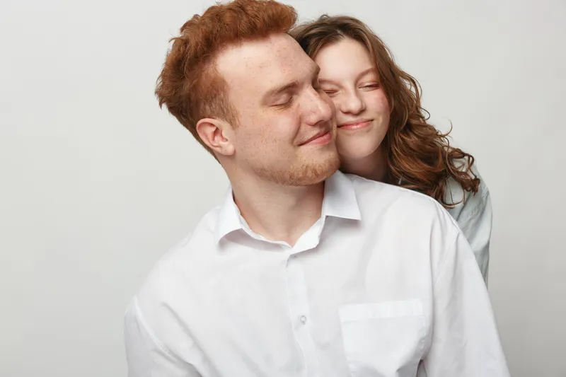 Happy young couple with closed eyes embraces gently, showing affection and love. Both wear white shirts against a plain light background. Concept of romance and intimacy.