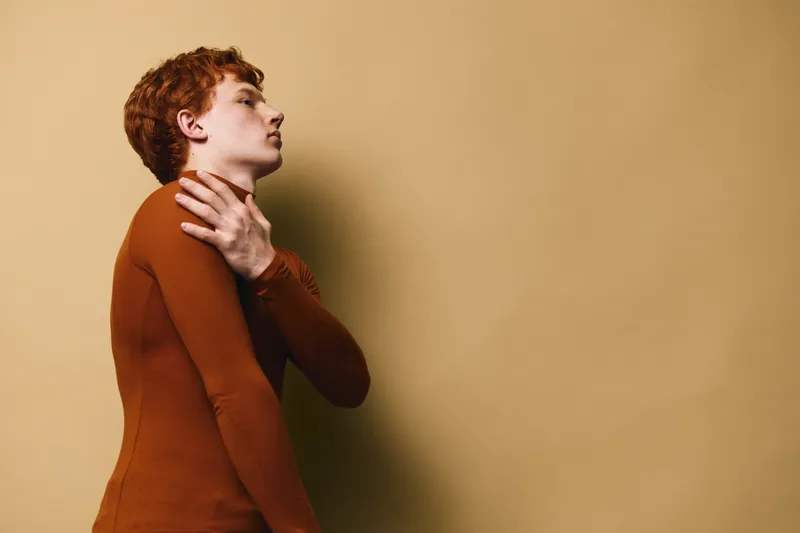 Young man with red hair wearing brown turtleneck touching his neck with hand while standing against beige background in side view portrait.