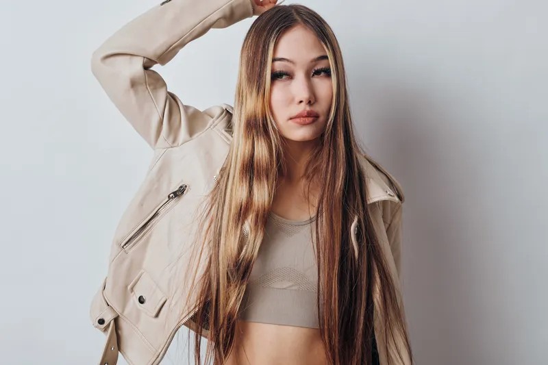 Young woman with long straight hair wearing beige leather jacket and crop top posing against white background with confident expression and casual style indoors.