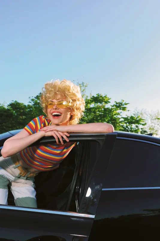 Joyful woman leaning from a black car window, outdoors on a sunny day, retro film color, candid smile, striped tee, breezy hair, vibrant natural backdrop