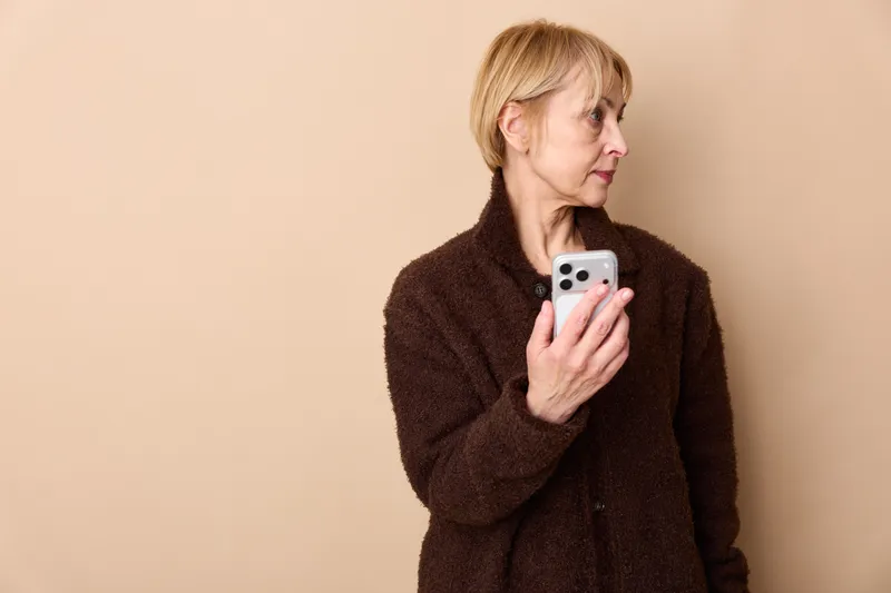 Elderly woman with smartphone, indoor portrait, side profile, brown coat, thoughtful expression, beige background, relaxed posture, soft communication, quiet moment