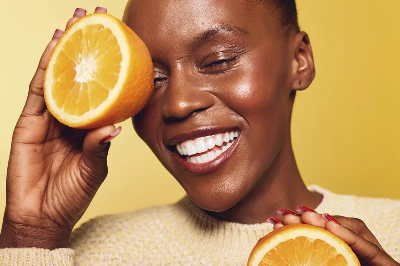 Happy African woman with dark skin smiling and holding two orange halves near her face, wearing a beige sweater on yellow background. Concept of healthy lifestyle and natural beauty.