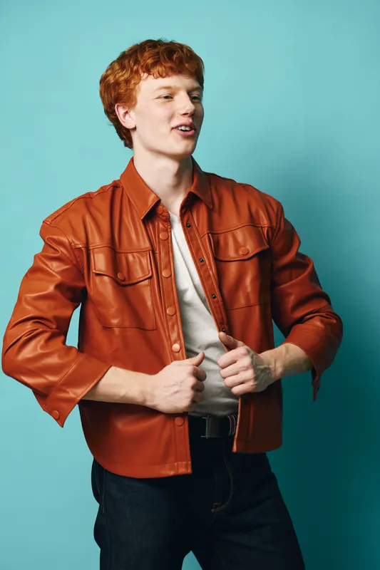 Young man with red hair wearing brown leather jacket and white shirt posing in front of blue background with confident expression and casual style.