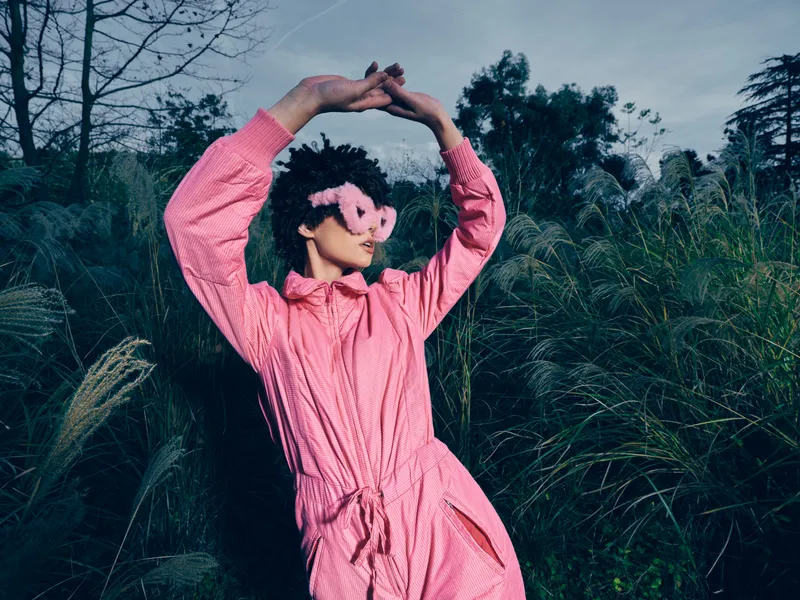 Woman in pink dress outdoors, blindfolded, striking a relaxed, graceful pose among tall grasses and lush greenery, conveying mystery, playfulness, and a nature inspired mood.