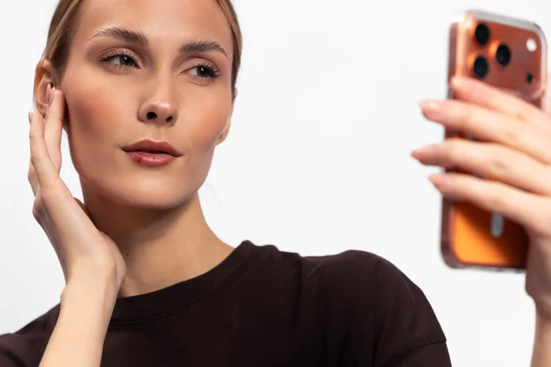 Young woman with clear skin and brown hair takes a selfie with a smartphone on white background. Concept of digital lifestyle, technology, beauty, and social media communication.