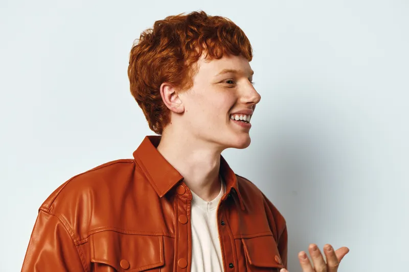 Young man with red hair wearing orange leather jacket smiling and speaking in casual style on white background. Portrait of happy male with expressive gesture.
