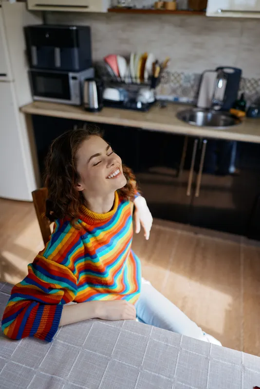 Cheerful woman indoors wearing a rainbow striped sweater, smiling at a kitchen table in a cozy home moment with natural light, relaxed lifestyle, casual vibe, and warm colors
