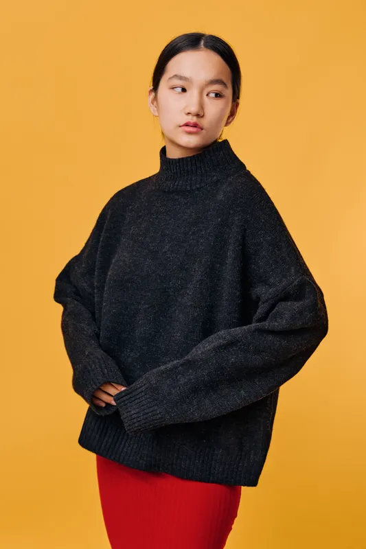 Woman wearing black oversized sweater and red skirt standing against yellow background looking sideways with thoughtful expression in casual fashion style portrait studio.