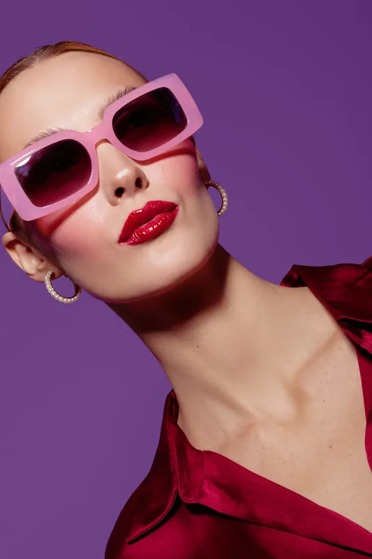 Stylish woman with pink sunglasses and red lipstick poses against purple background wearing red satin blouse and hoop earrings. Fashion portrait with bold makeup and accessories.