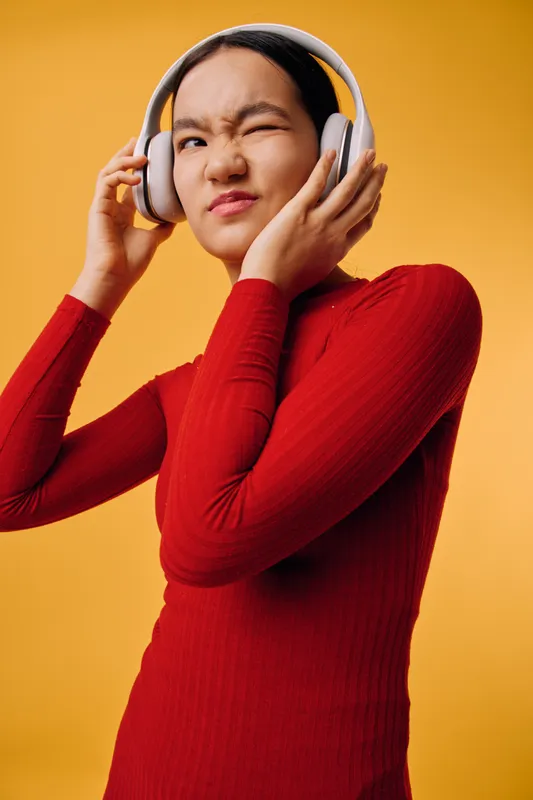 Young woman in red sweater listens to music with headphones and expressive face on orange background. Casual portrait with emotion, technology, entertainment and style in studio lighting.