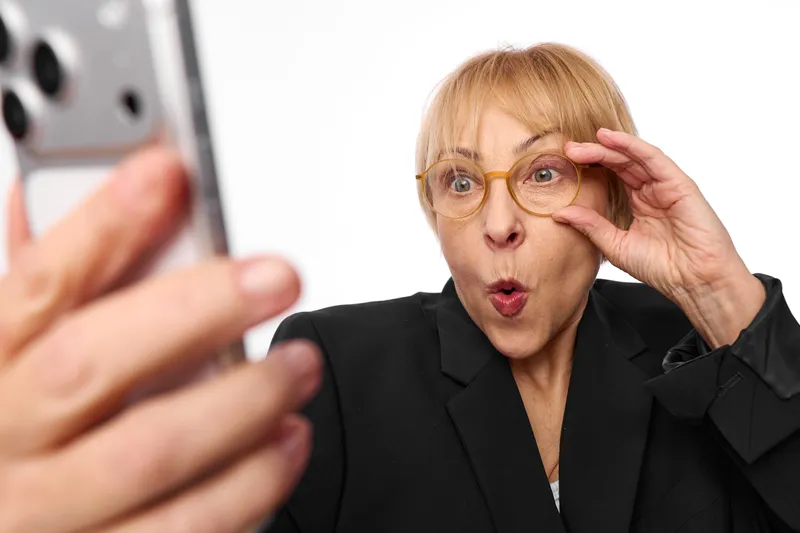 Woman with eyeglasses looks surprised while adjusting frames, close up portrait in office. Hand holds smartphone, concept of technology, communication, alertness.