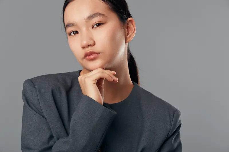 Portrait of young Asian woman with dark hair wearing gray jacket looking at camera with thoughtful expression on plain gray background. Female model with natural makeup and confident pose.