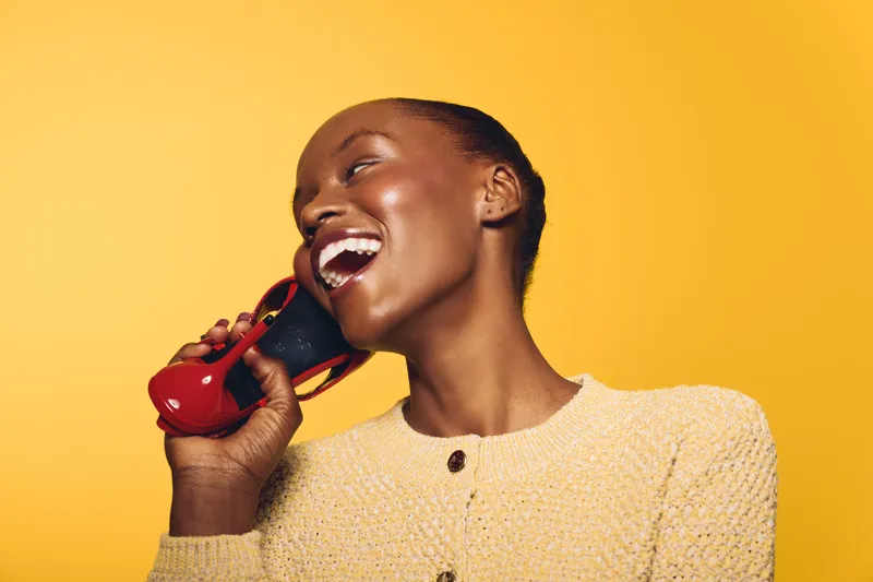 Smiling black woman with dark skin wearing a yellow sweater holds a red vintage telephone receiver near her mouth. Bright yellow background creates a joyful and vibrant atmosphere.