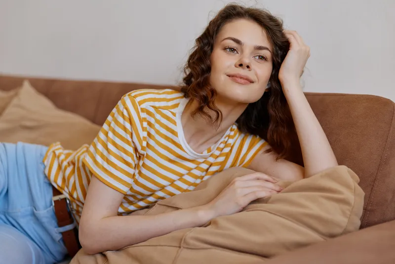 Young woman relaxes on a cozy couch at home, wearing a yellow striped shirt and jeans, with a calm smile, creating a warm casual lifestyle moment on a comfortable living room setting