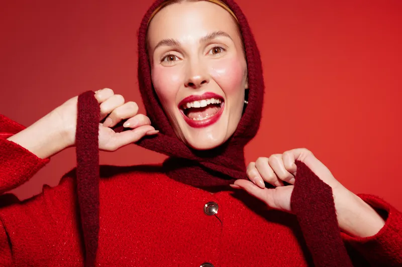 Smiling woman in red sweater and hood pulls hood strings on red background. Bright makeup and joyful expression create festive and cheerful mood. Concept for happiness and winter fashion.