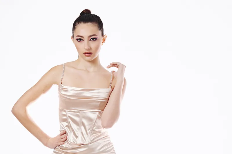 Young woman in a shiny satin dress poses confidently against a white background. Her hair is styled, makeup bold, concept for beauty, fashion, and creative portrait lifestyle.