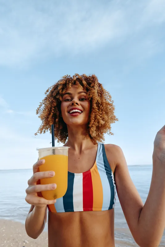 Woman on beach smiles while holding an iced drink with a straw, wearing a striped swimsuit. Ocean horizon and clear blue sky suggest summer vacation. Curly hair in the breeze near water.